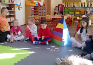 Dzieci siedzące w półkolu. Obok "piramida żywienia" (przestrzenny kształt trójkąta) z oznaczonymi kolorowymi piętrami.